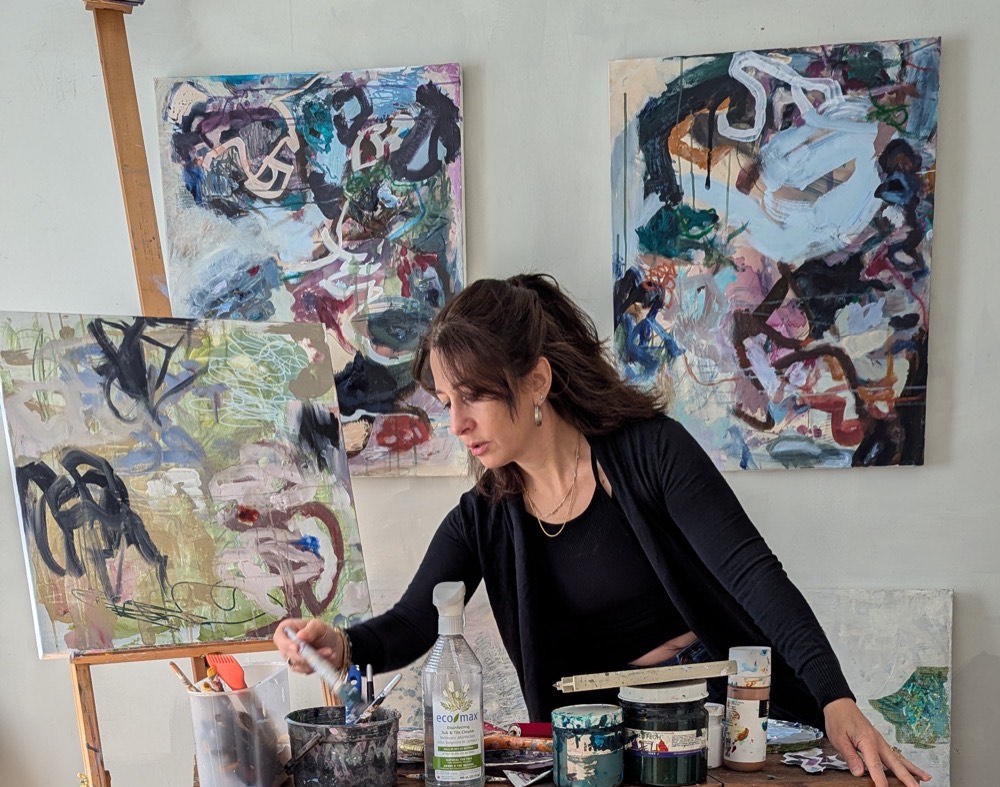 Sari Zon at work in her studio, reaching for a brush, surrounded by her abstract paintings